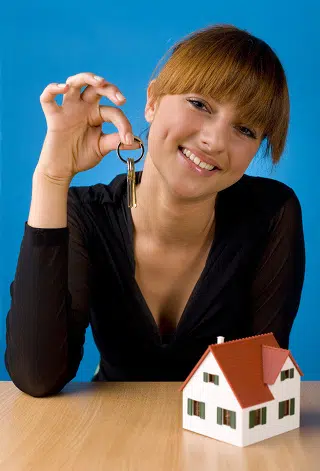Beautiful woman sitting at desk in front of house miniature. Holding keys. Smiling and looking at camera. Front view, blue background